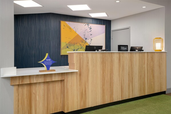 Uma receção de hotel com design moderno, apresentando um balcão de madeira, uma grande pintura abstrata na parede e uma placa com o nome do hotel.