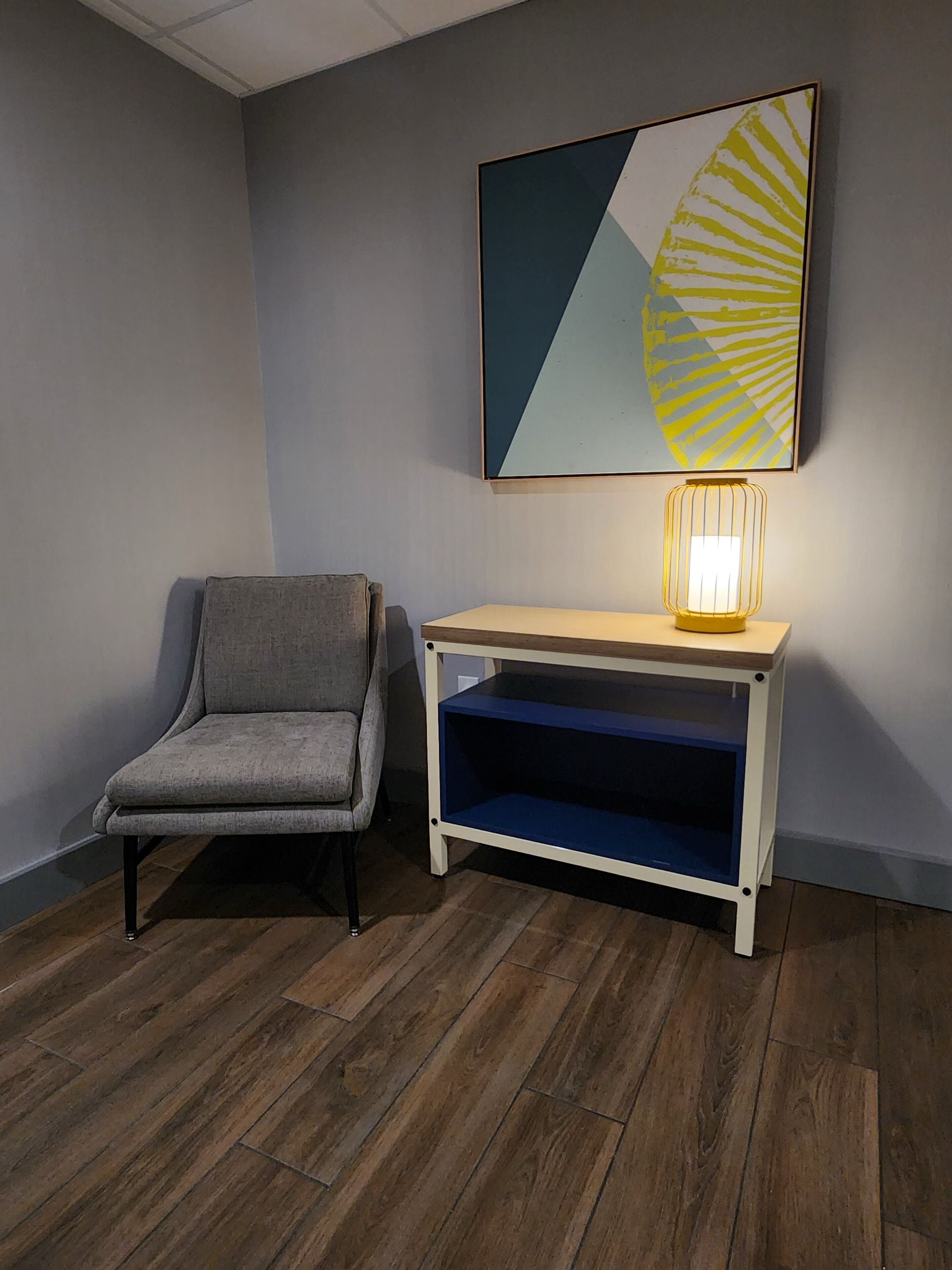 A corner room with a grey upholstered chair, a wooden console table with a blue shelf, and a wall-mounted abstract painting.