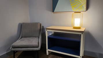 A corner room with a grey upholstered chair, a wooden console table with a blue shelf, and a wall-mounted abstract painting.