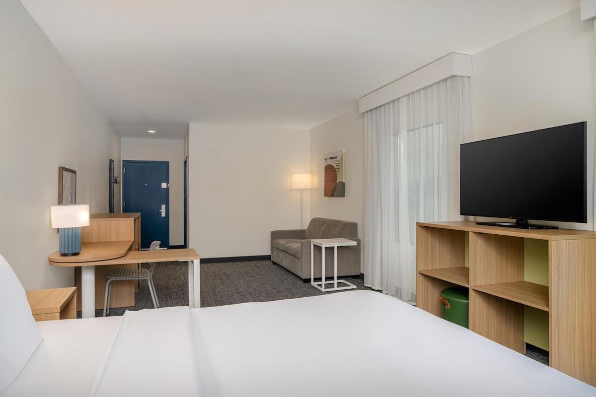 Ett hotellrum med en säng, ett skrivbord med en lampa, en tv, en soffa och en dörr.