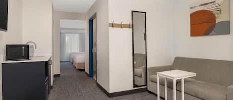 A hotel room with a sofa, a small table, a mirror, a coat rack, and a television on a stand.