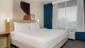 A hotel room with a large bed, a bedside table, a wall-mounted artwork, a window with sheer curtains, and a wall heater.