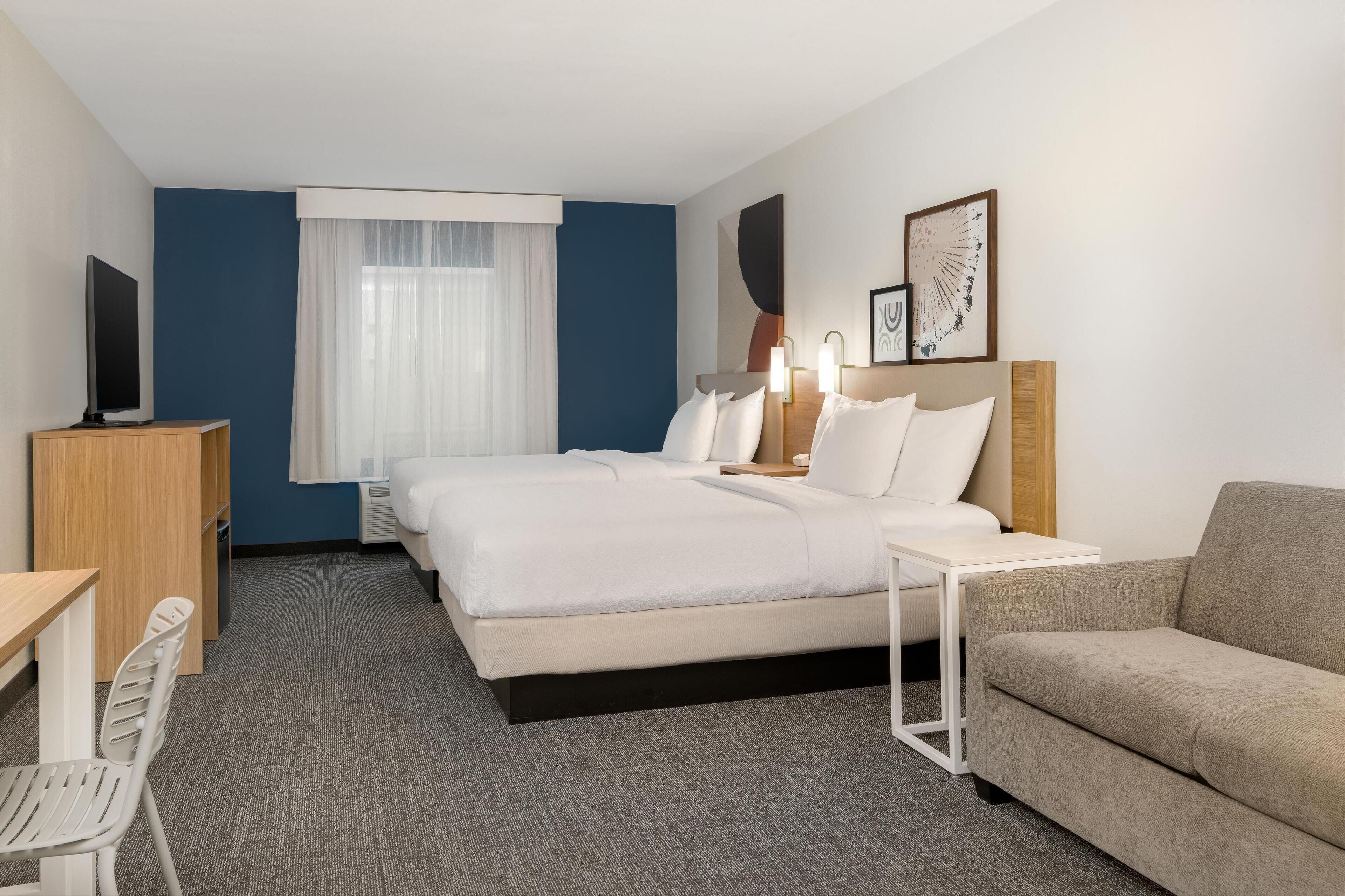 A hotel room with a bed, a sofa, a desk with a chair, a television, and a window with curtains.