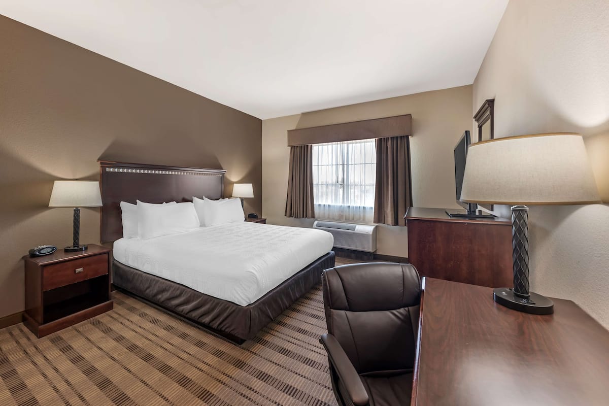 A hotel room with a large bed, a desk, a chair, a nightstand, and a window with curtains.