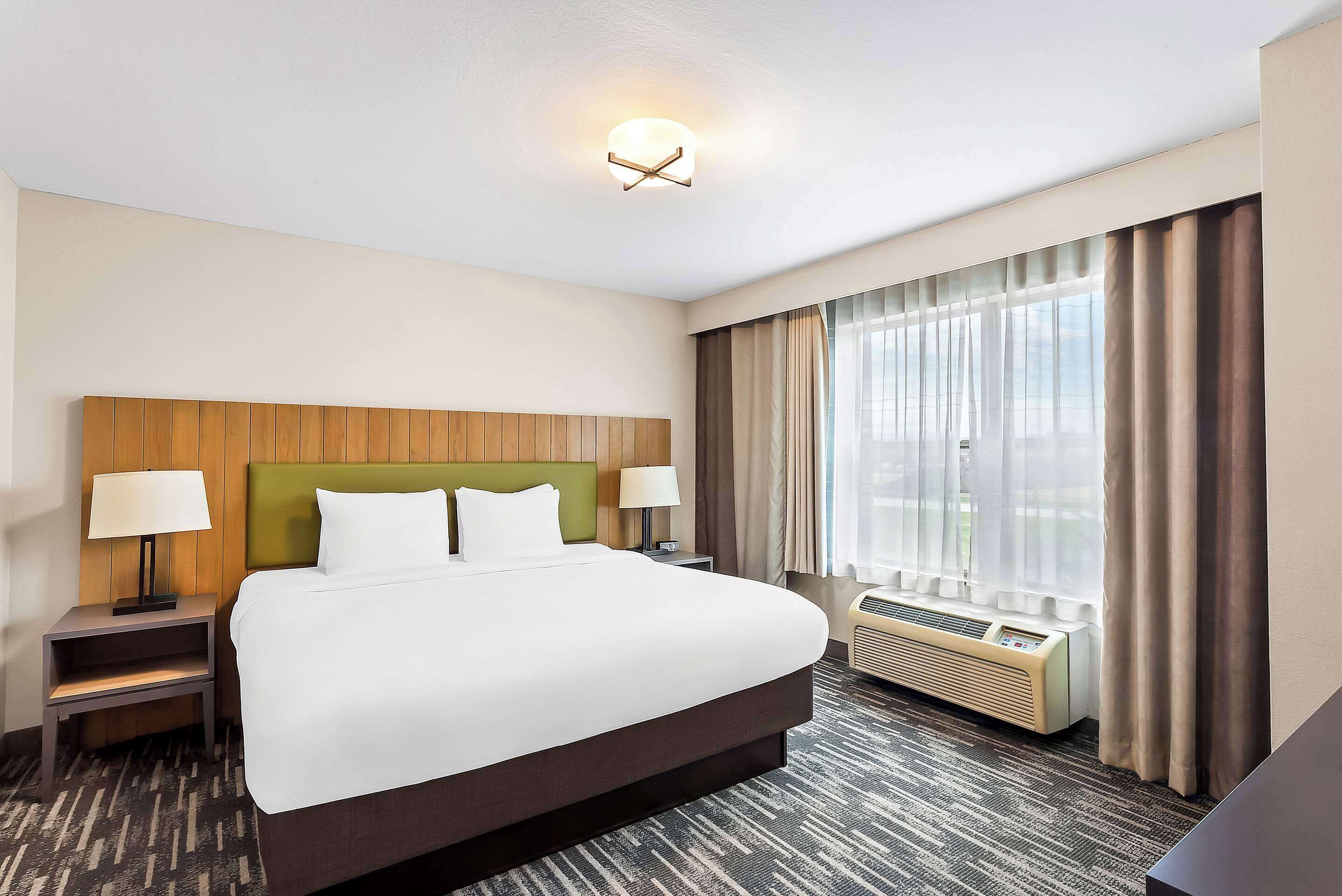 Una habitación de hotel con una cama grande, dos mesitas de noche con lámparas, un cabecero, una ventana con cortinas y un equipo de aire acondicionado.