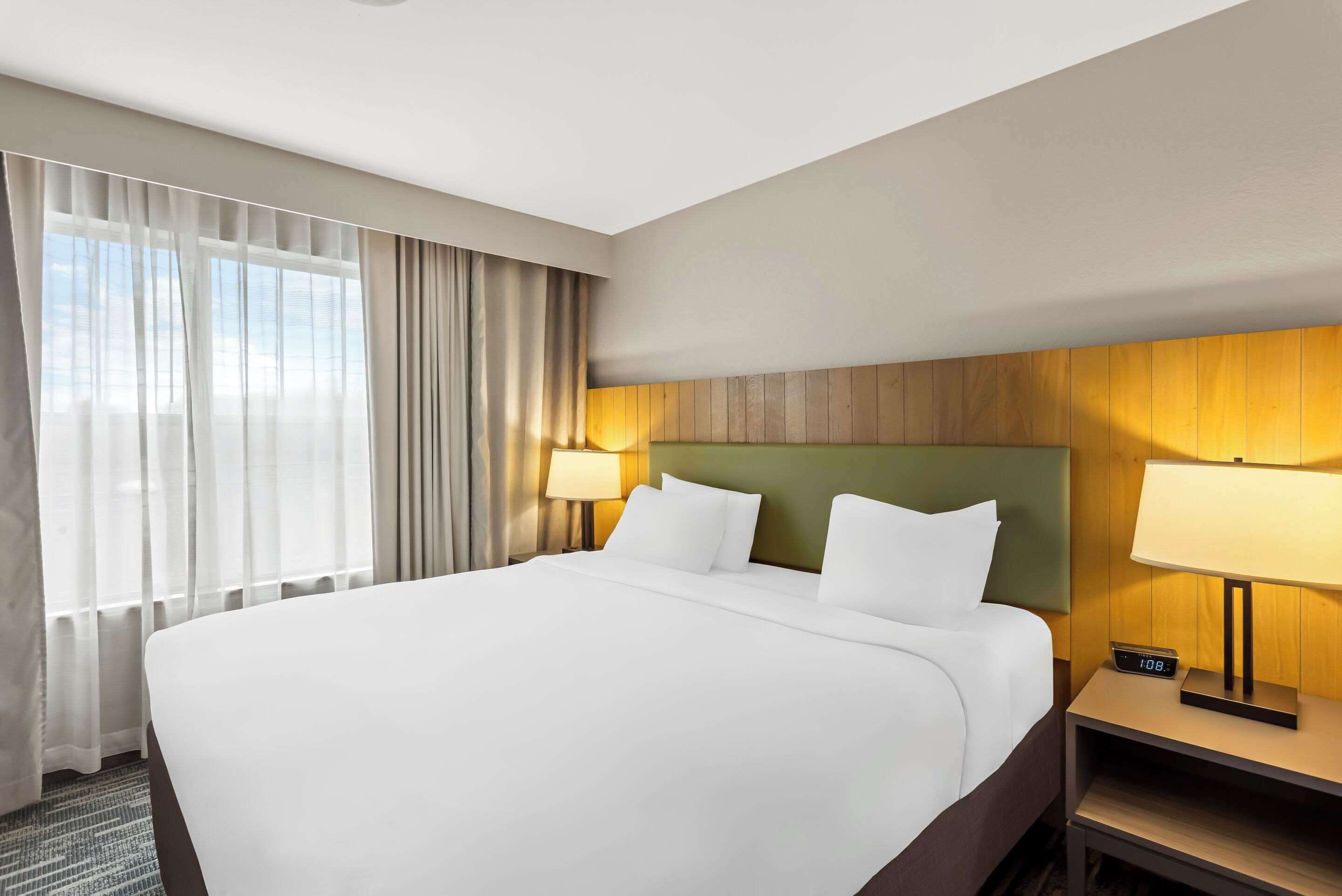 Habitación de hotel con una cama grande, dos lámparas de noche, una mesita de noche y vistas al exterior a través de cortinas translúcidas.