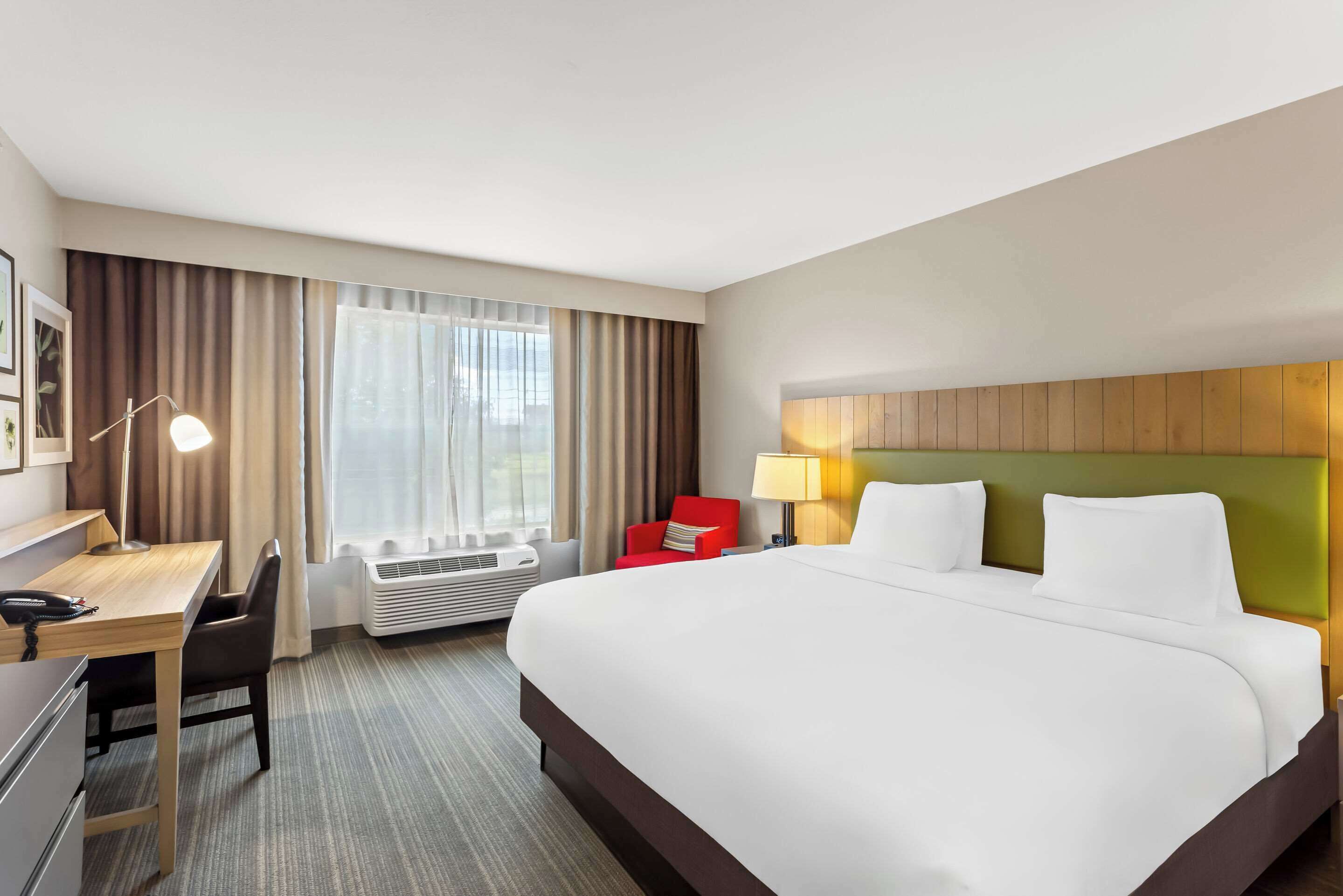 Une chambre d’hôtel avec un grand lit, un bureau avec une lampe, une chaise rouge, un appareil de climatisation et une fenêtre avec des rideaux.