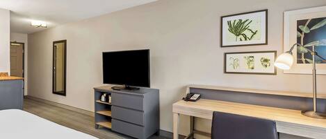A hotel room with a bed, a desk with a chair, a television, a desk lamp, a mirror, and framed botanical artwork on the wall.