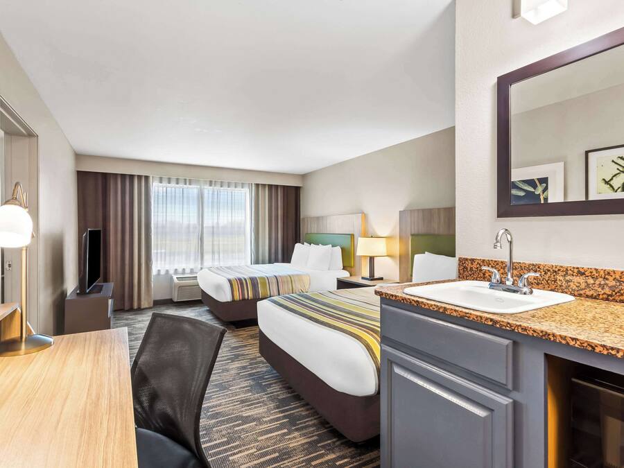 Habitación de hotel con dos camas, un escritorio, una silla, un televisor, un espejo grande, un lavamanos y una ventana con cortinas.