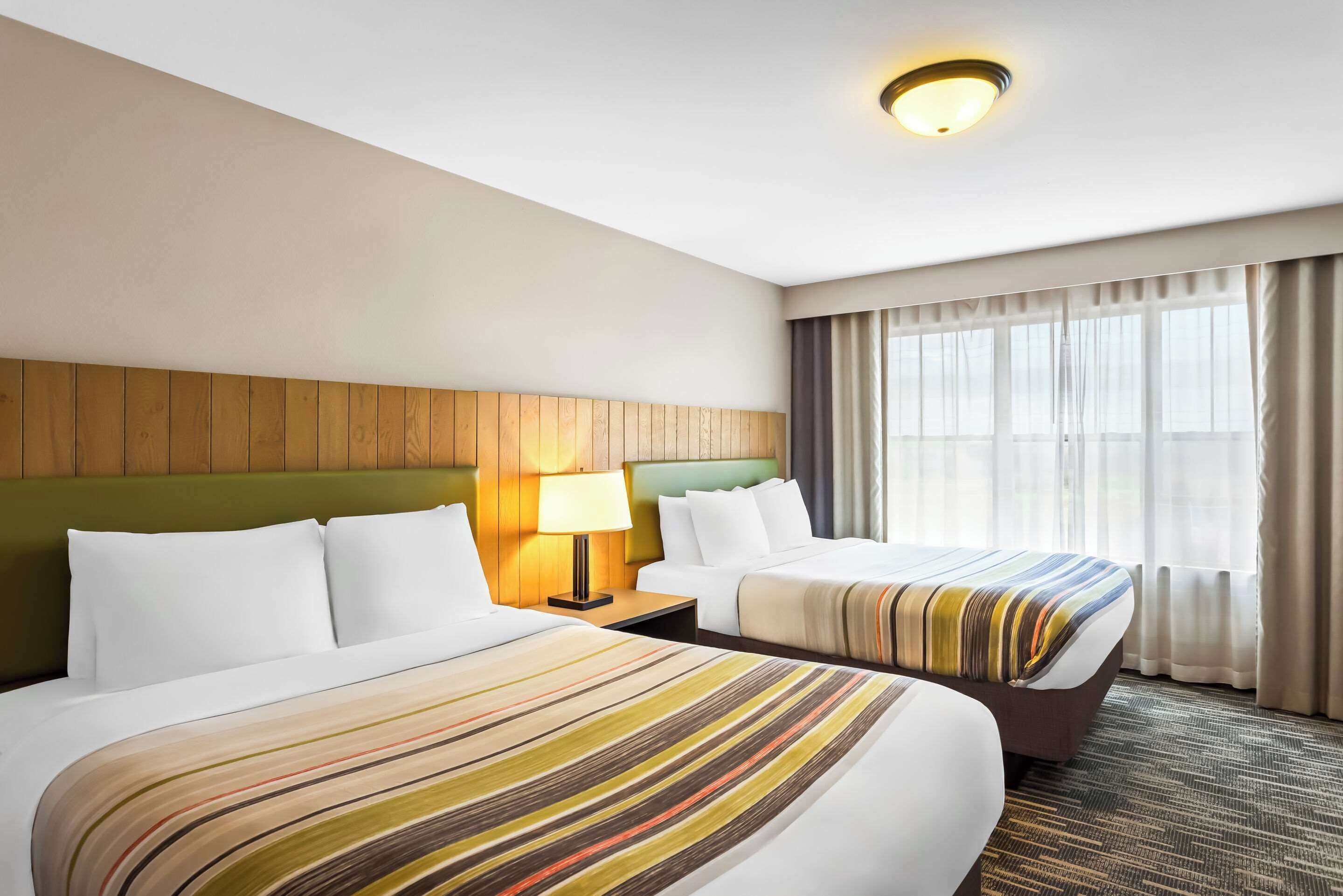 Habitación de hotel con dos camas, cabecera de madera, mesita de noche con lámpara y una ventana grande con cortinas.