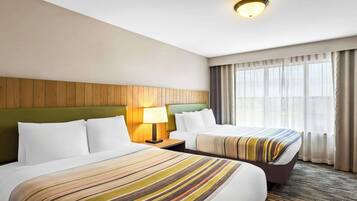 Habitación de hotel con dos camas, cabecera de madera, mesita de noche con lámpara y una ventana grande con cortinas.