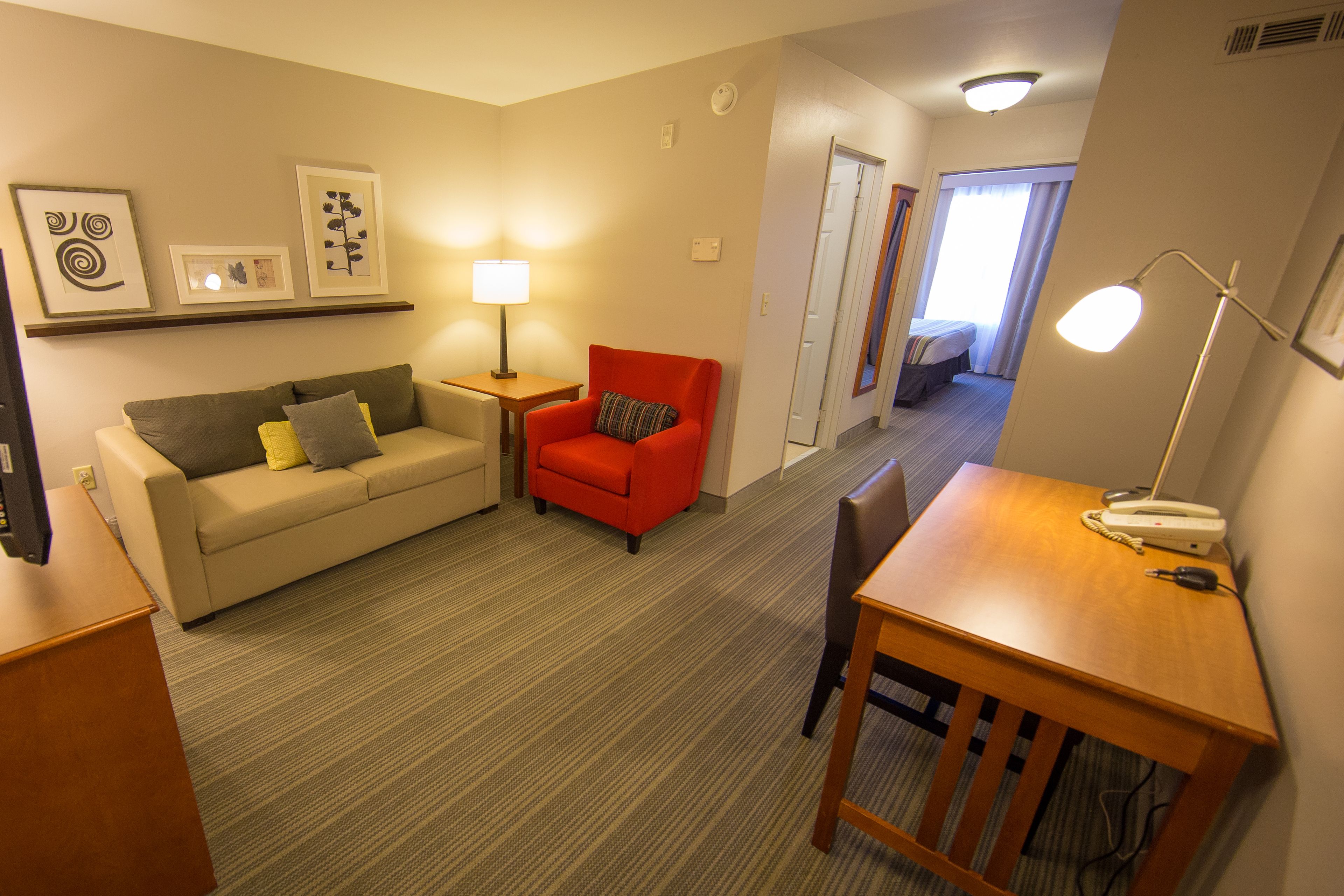 A hotel room with a sofa, a red armchair, a wooden table, and a desk. There are framed pictures on the wall, a lamp on the table, and a door leading to a balcony.