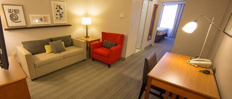 A hotel room with a sofa, a red armchair, a wooden table, and a desk. There are framed pictures on the wall, a lamp on the table, and a door leading to a balcony.