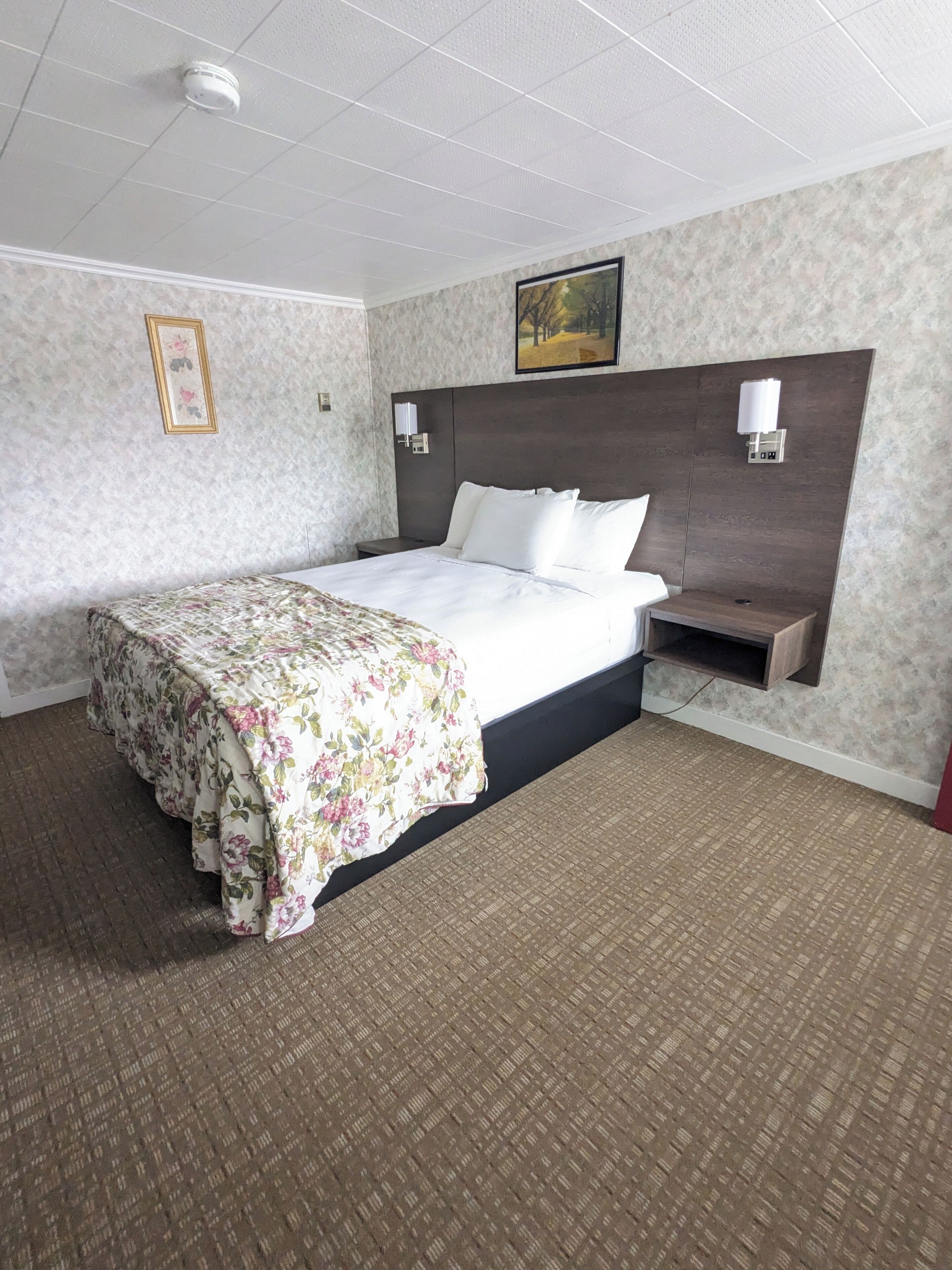 Habitación de hotel con una cama grande, un cabecero con luces empotradas en la pared, una pequeña estantería y un detector de humo instalado en el techo.