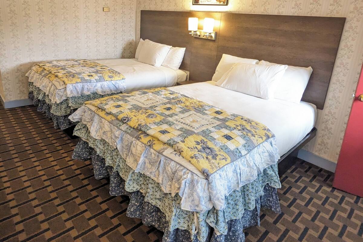 Hotellihuone, jossa on kaksi sänkyä, kukkakuviotapetti, kuvioitu lattia ja taulu sängynpäädyn yläpuolella.