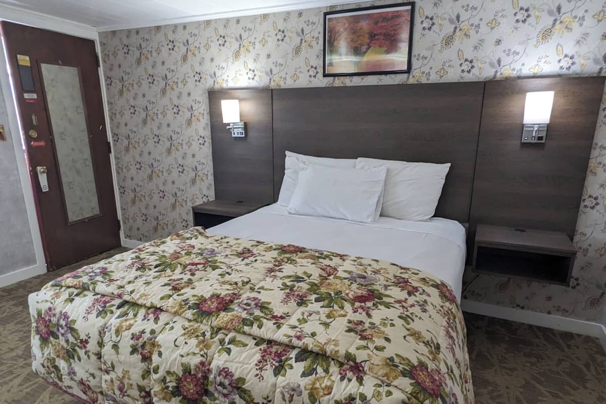 Hotellihuone, jossa on kukkakuviollinen päiväpeite, puinen pääty, kaksi yöpöytää valaisimineen ja kukkakuviotapetti.