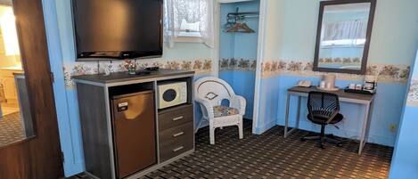 Ein kleiner Raum mit Fernseher, einem Schreibtisch mit Stuhl, einem Kleiderschrank und einem gemusterten Boden.