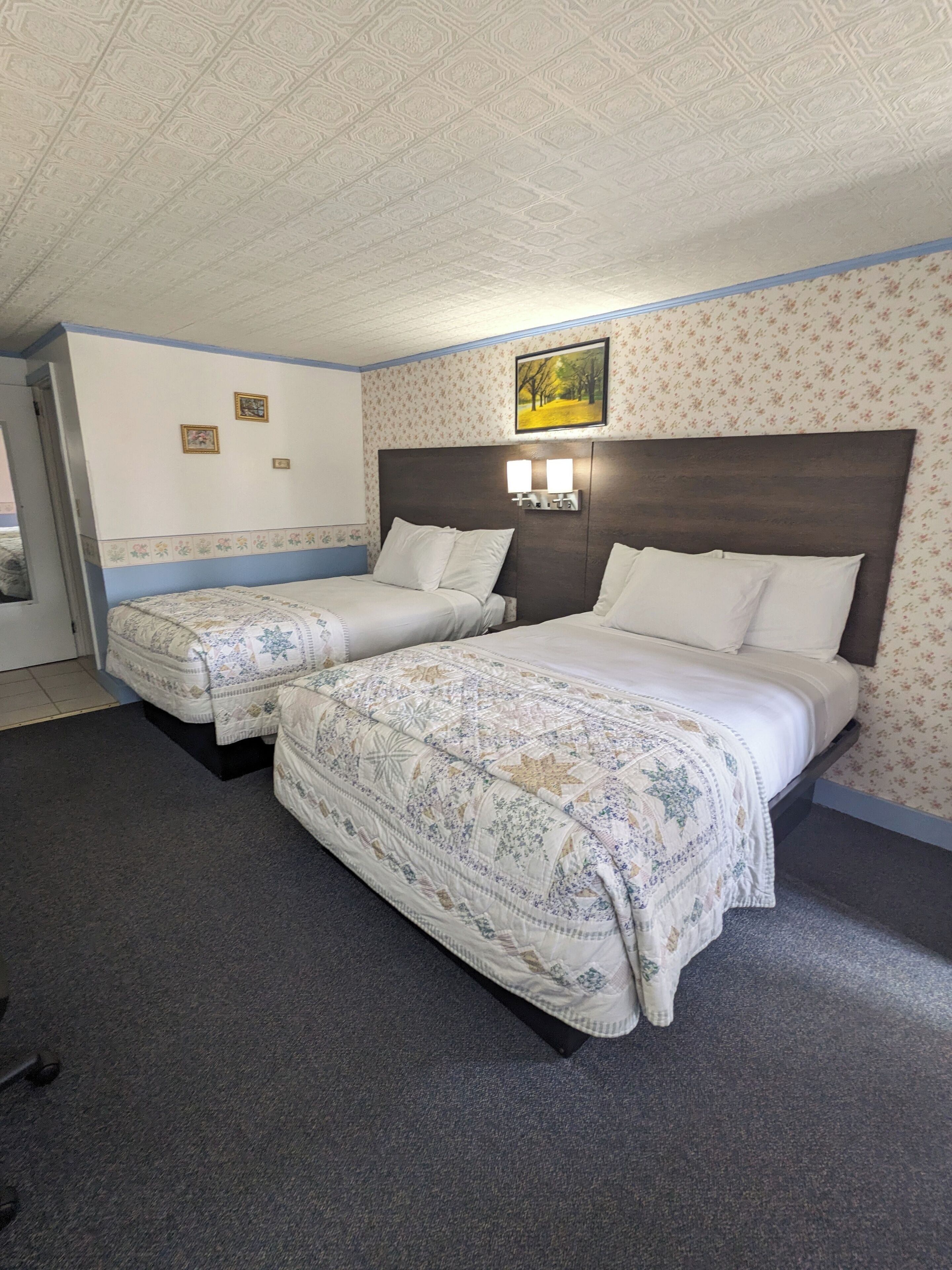 A hotel room with two beds, a headboard with wall-mounted lights, floral wallpaper, and a framed picture on the wall.
