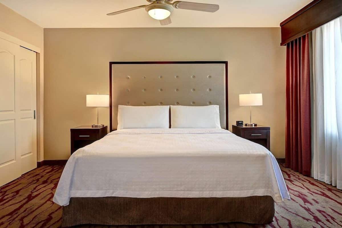 A hotel room with a large bed, two bedside tables, a ceiling fan, and a window with curtains.