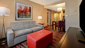 A hotel room with a grey sofa, red ottoman, flat-screen TV, and a painting of mountains.
