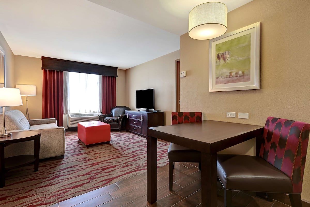 Une chambre d’hôtel avec un canapé, une table à manger, un pouf rouge, une télévision et un tableau au mur.