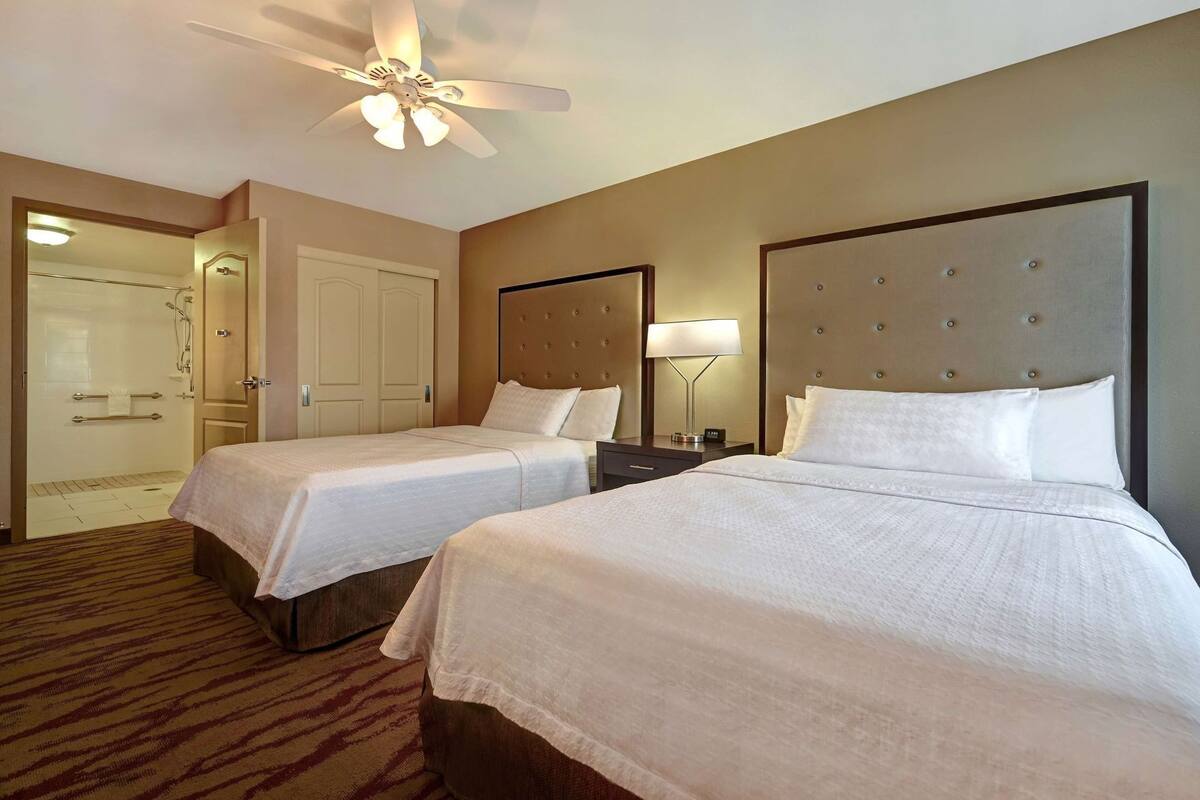 A hotel room with two beds, a ceiling fan, a nightstand with a lamp, and a bathroom with a shower and towel rack.