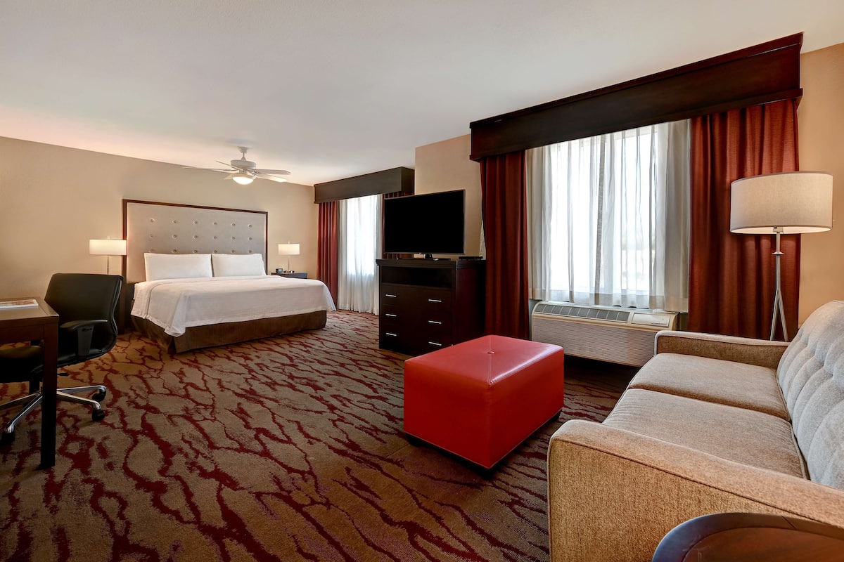 Une chambre d’hôtel comprenant un lit, un bureau, une télévision, un pouf rouge, un canapé et une grande fenêtre avec des rideaux.