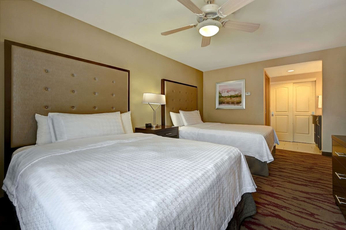 Une chambre d’hôtel avec deux lits, un ventilateur de plafond, une table de chevet avec une lampe, un tableau au mur et une porte donnant sur une autre pièce.