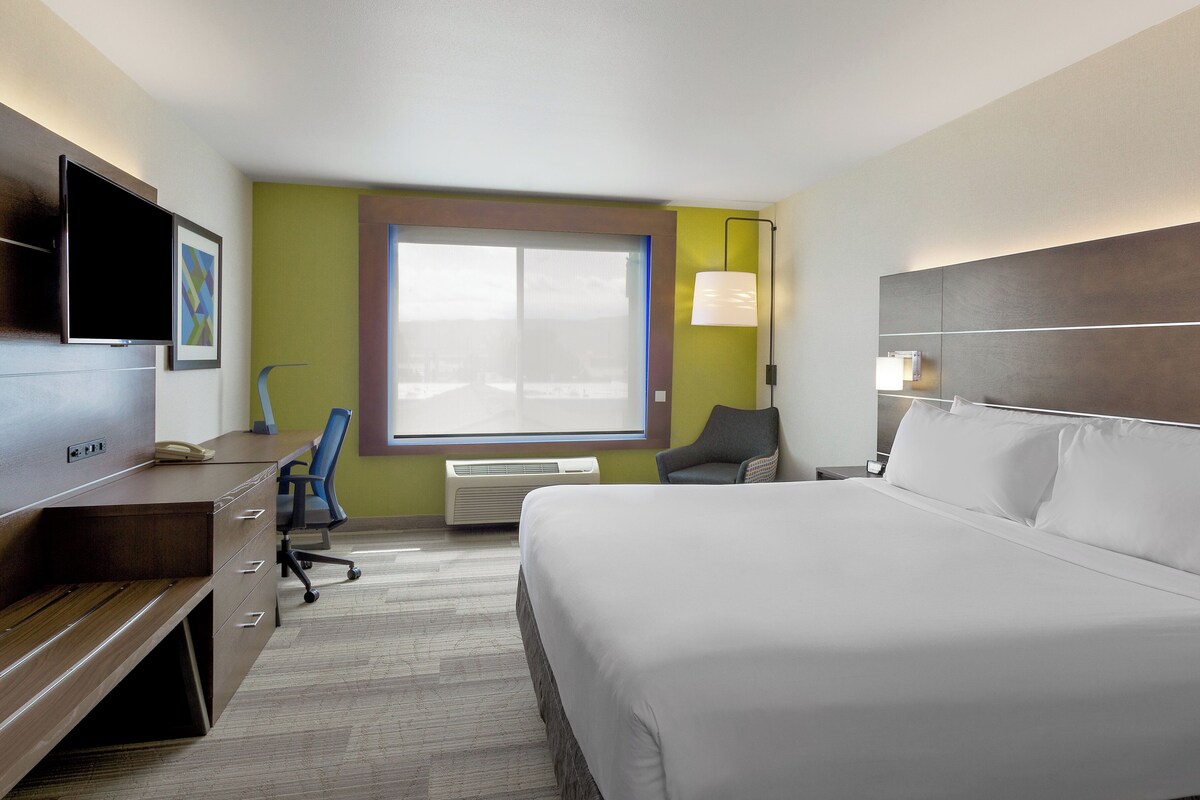 A hotel room with a bed, desk, chair, TV, air conditioning unit, and a window with blinds.