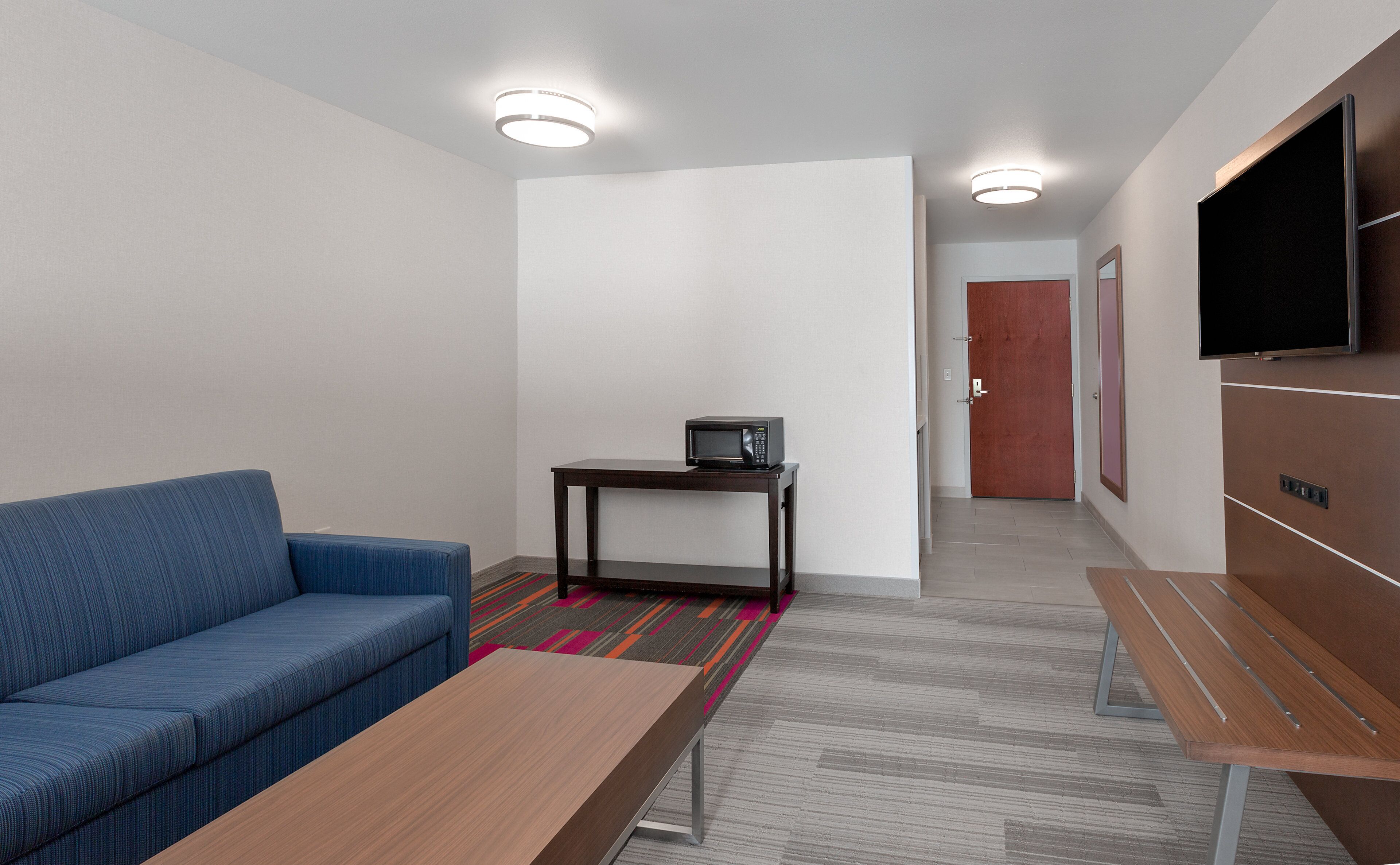A modern hotel room with a blue sofa, a wooden table, a small table with a microwave, a TV mounted on the wall, and a door leading to another room.