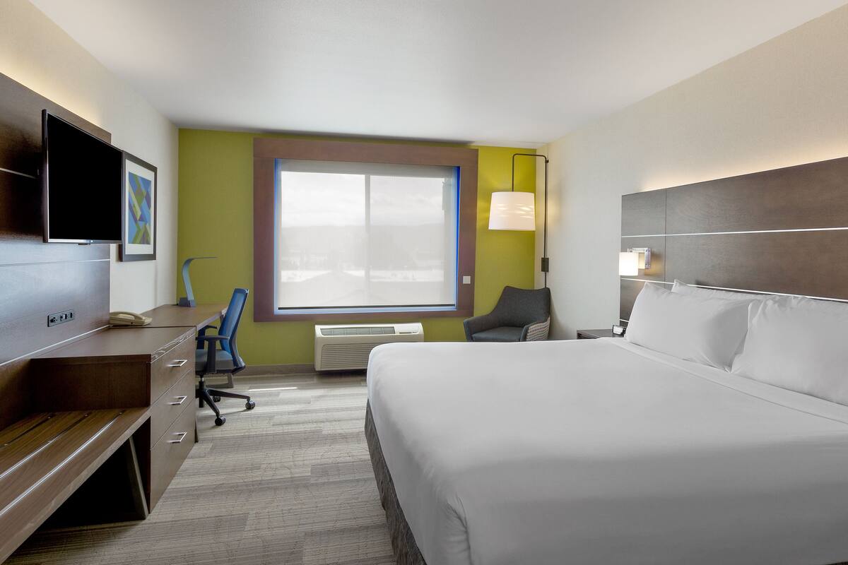 A hotel room with a bed, desk, chair, TV, air conditioning unit, and a window with blinds.