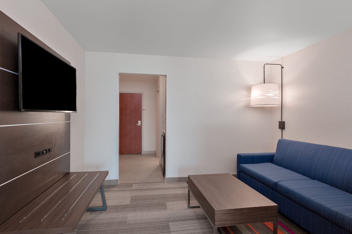 A modern hotel room with a flat-screen TV mounted on the wall, a blue sofa, a wooden coffee table, and a floor lamp.