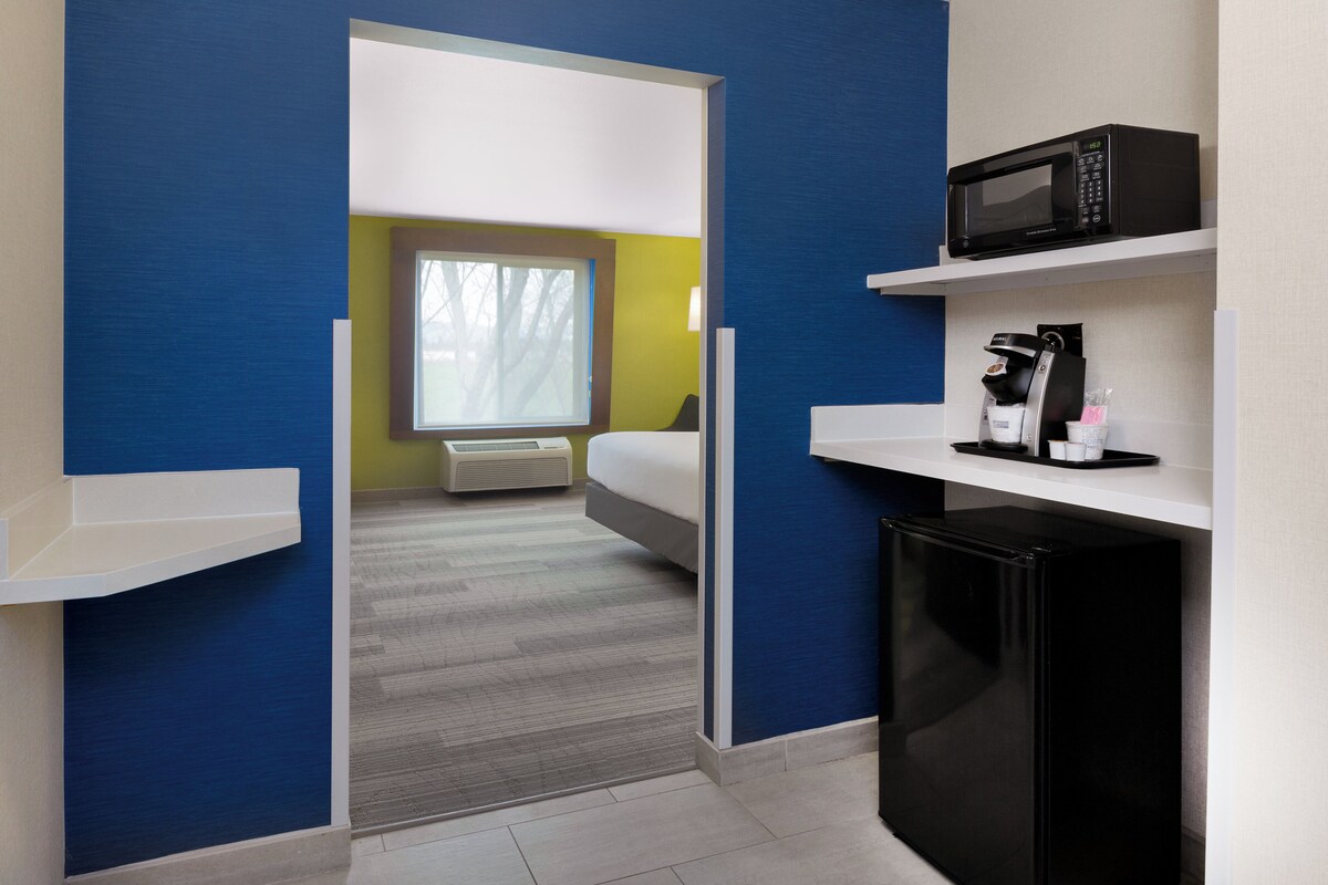 A modern hotel room with a bed, a microwave, a coffee maker, and a small refrigerator.