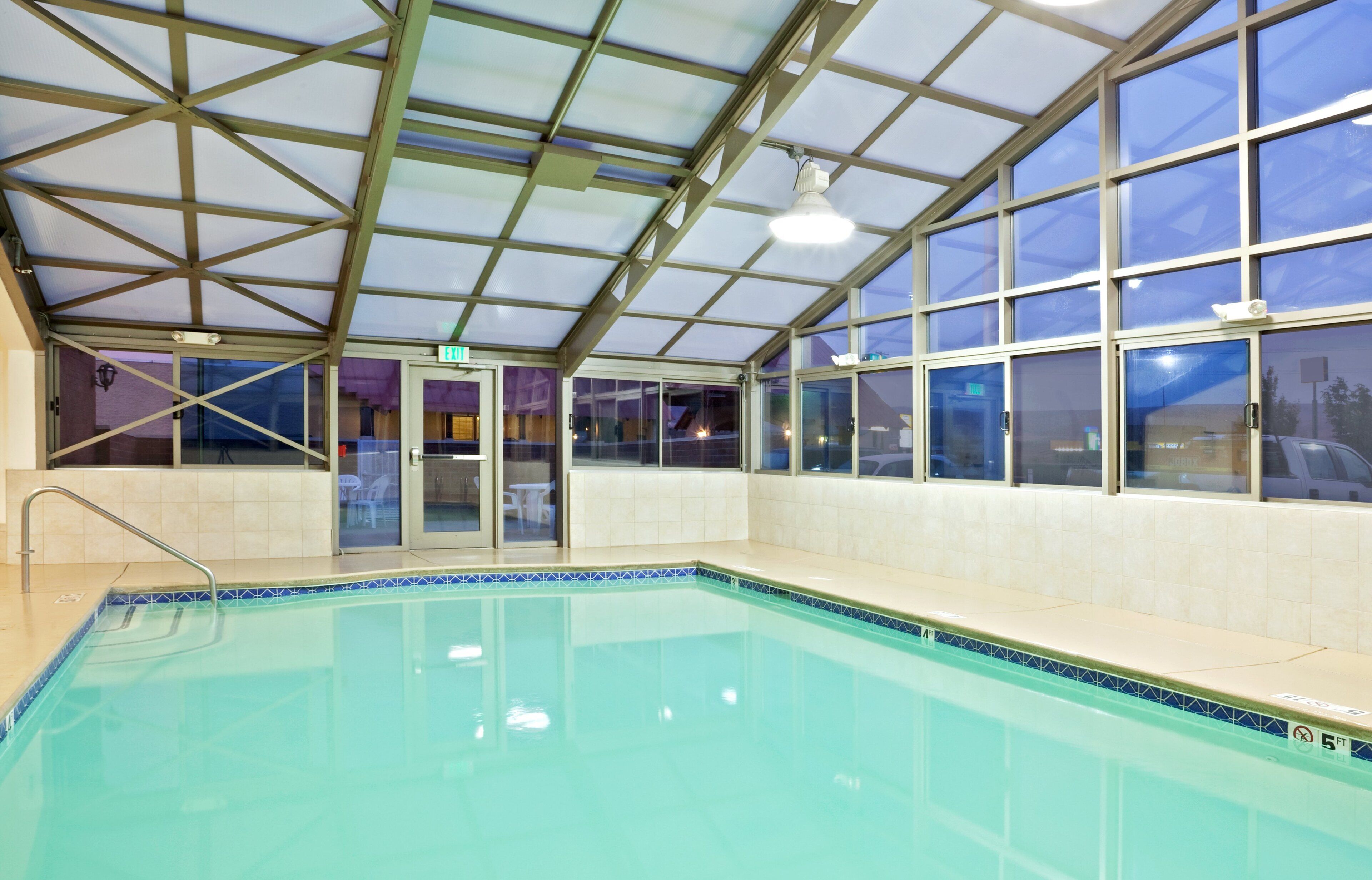 An indoor swimming pool with clear glass walls, a metal roof structure, and a tiled floor.