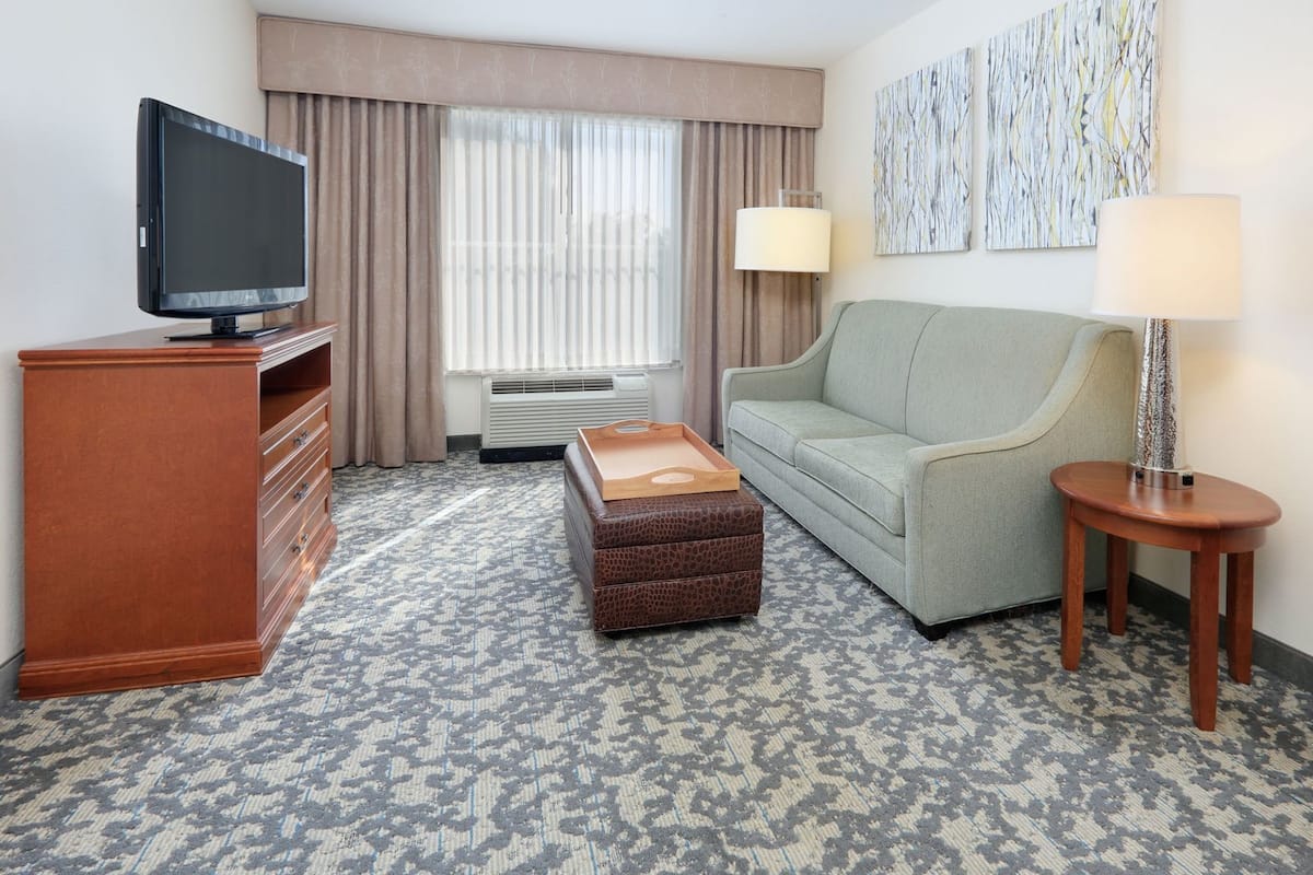 Ein Hotelzimmer mit Flachbildfernseher, einer Couch, einem Holznachttisch, einer Lampe, einem gemusterten Teppich und einem Fenster mit Vorhängen.