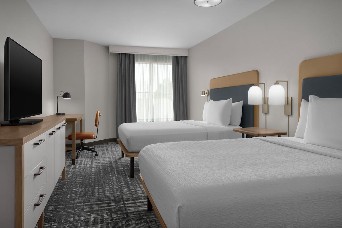 Ein Hotelzimmer mit zwei Betten, einem Schreibtisch mit Fernseher, einem Sessel und einem Fenster mit Vorhängen.