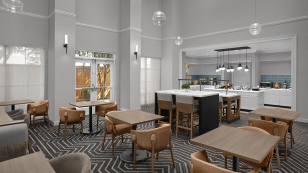 Un hall d’hôtel moderne, doté de hauts plafonds, d’un espace central avec des sièges et d’un coin cuisine visible au fond.