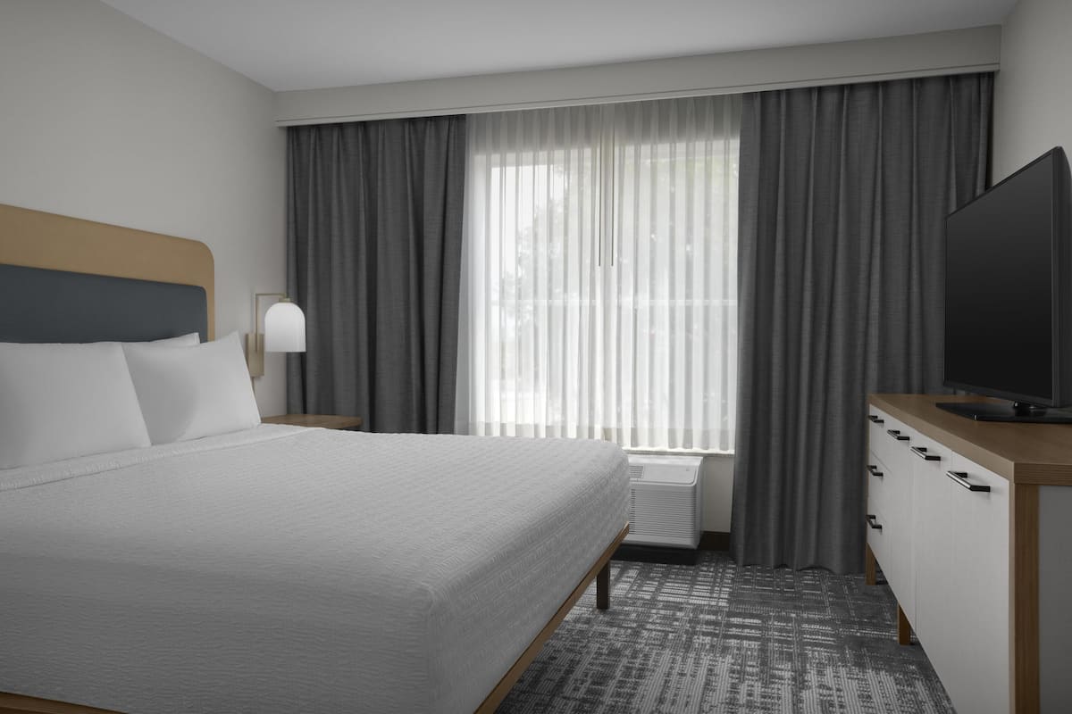 Ein Hotelzimmer mit einem Bett, einem Fernseher, einem Schreibtisch mit Schubladen, einer Lampe und einem Fenster mit Vorhängen.