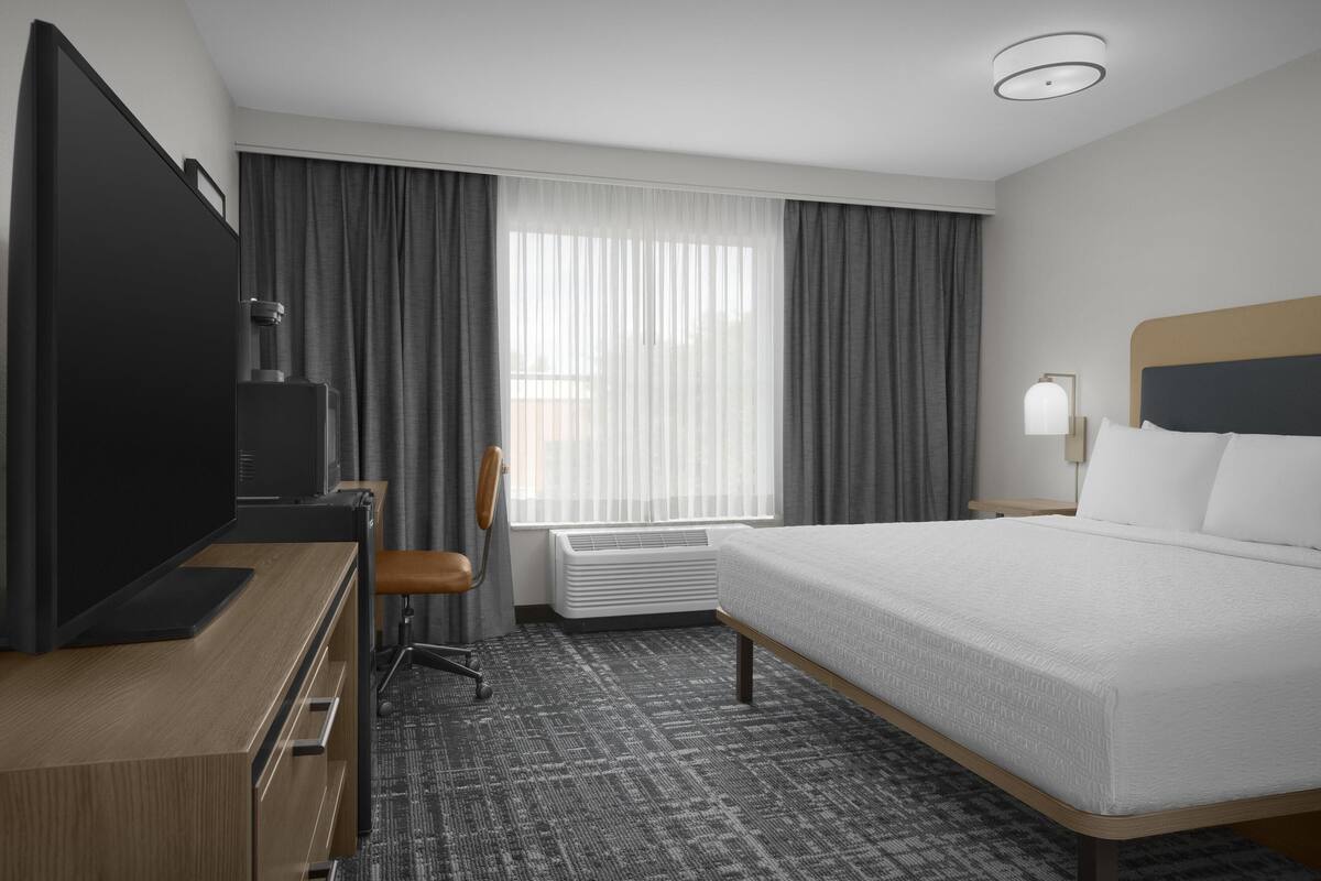 Ein Hotelzimmer mit einem Bett, einem Schreibtisch mit Fernseher, einem Sessel, einem Fenster mit Vorhängen und einer Wandleuchte.