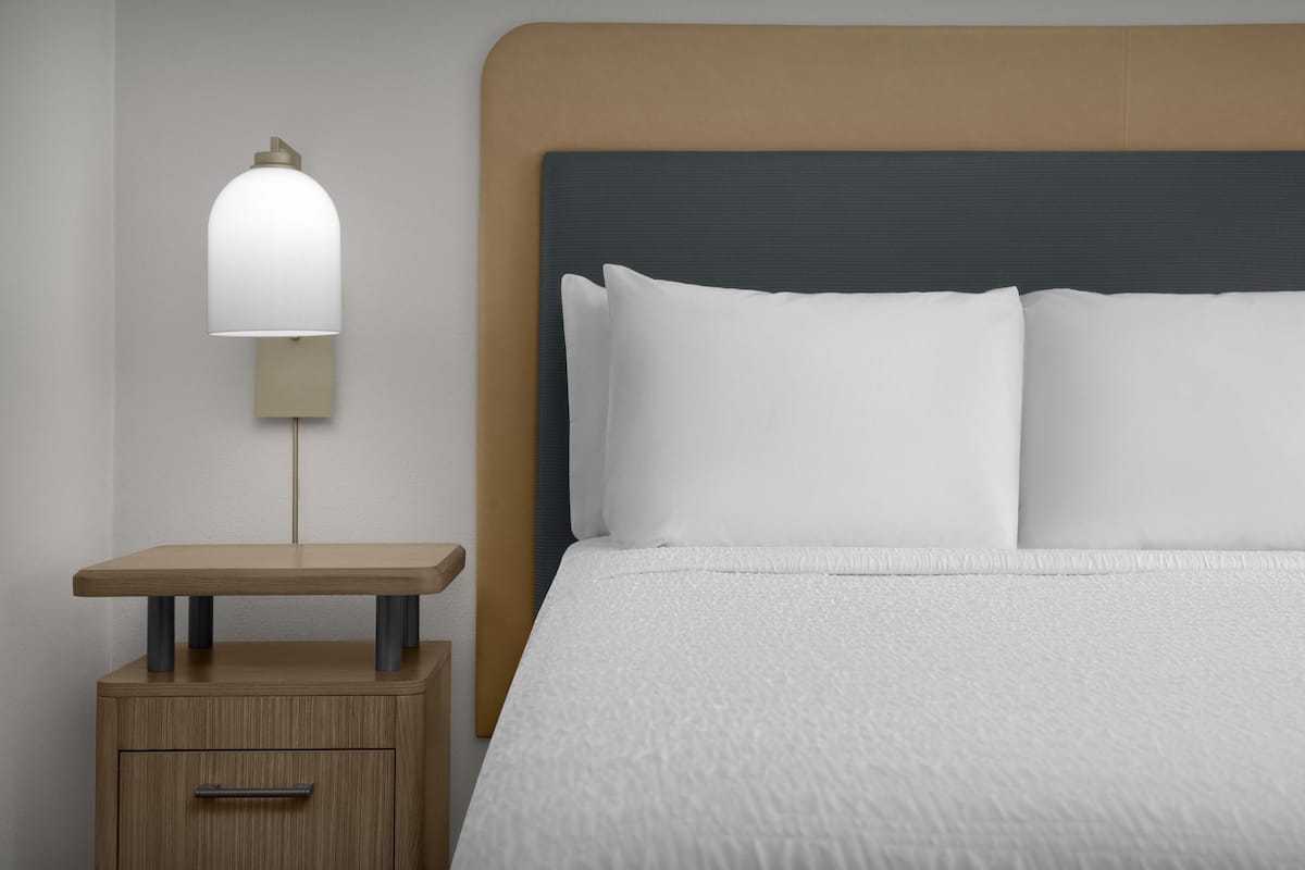 Ein ordentlich bezogenes Bett mit weißen Leinen, ein Nachttisch mit Lampe und eine Kommode.