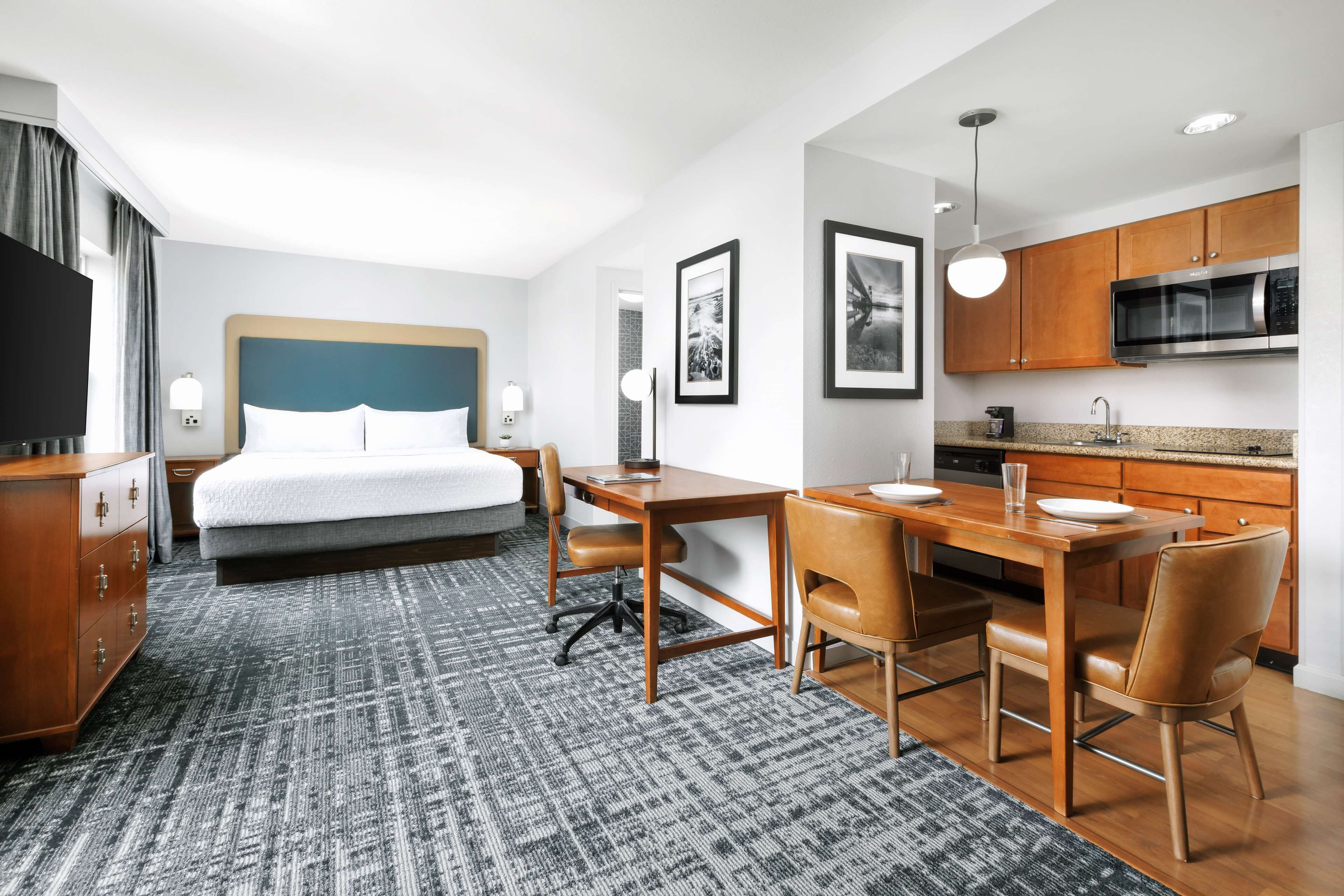 A hotel room with a bed, desk, chairs, a kitchen area, and a TV.