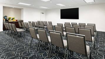 A conference room with rows of chairs, a large screen, and a kitchenette area with a coffee machine and fruit.