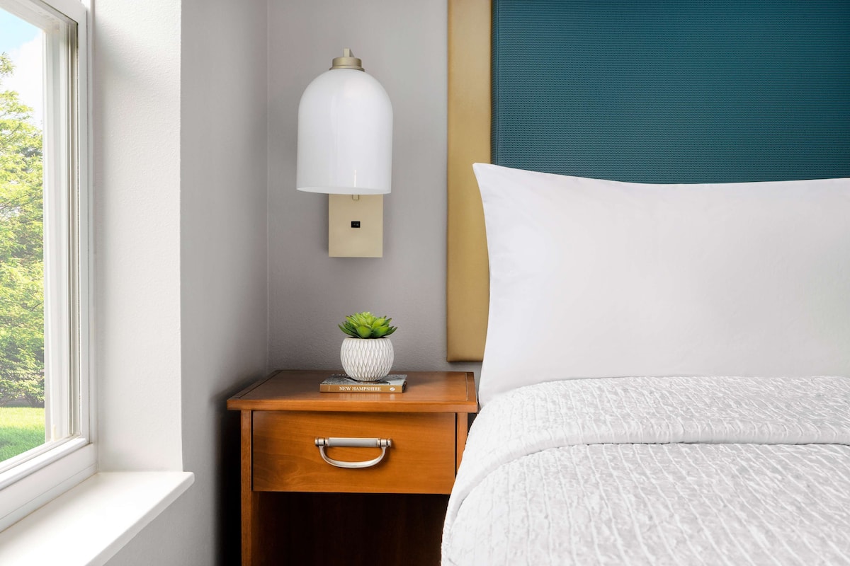 A neatly made bed with a white pillow and a textured white duvet. A wooden nightstand with a small potted plant and a book titled "New Hampshire" is beside the bed. A wall-mounted light fixture is above the nightstand.
