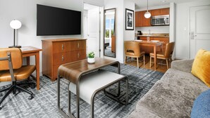 A modern living room with a sofa, a coffee table, a TV, a desk, and a kitchen area with a microwave and cabinets. - Homewood Suites by Hilton Portsmouth (Portsmouth)