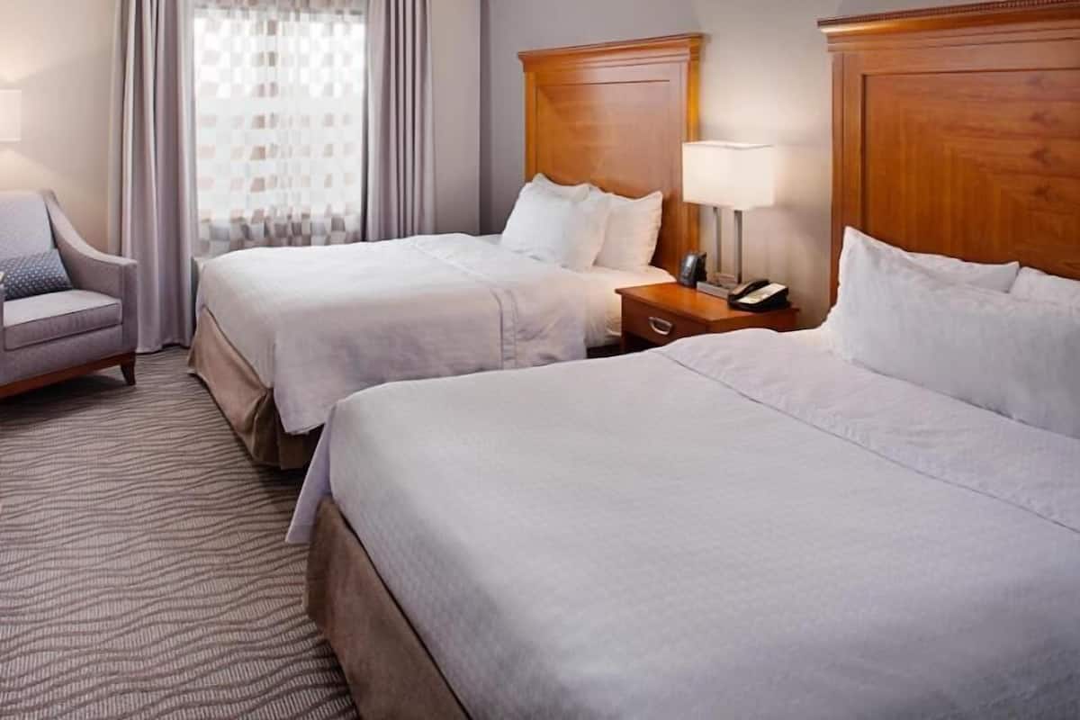 A hotel room with two beds, a chair, a nightstand with a lamp, and a window with curtains.