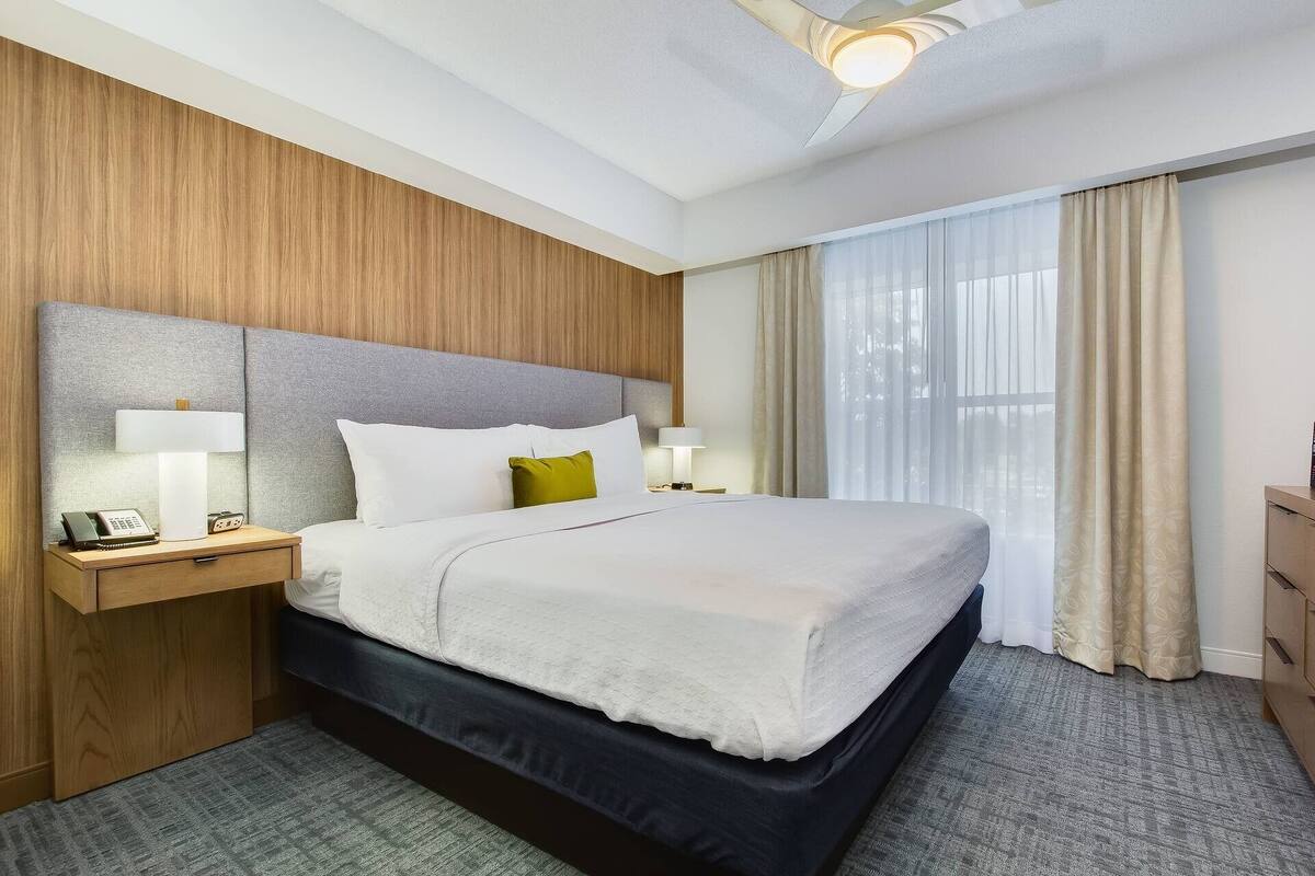 Una camera d'albergo con un letto grande, due comodini con lampade, una testiera in legno, una finestra con tende e una luce a soffitto.