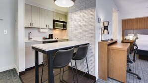 A compact hotel room with a kitchenette, a small dining area, a bed, and a bedside table with a lamp. - Homewood Suites by Hilton Louisville-East (Louisville)