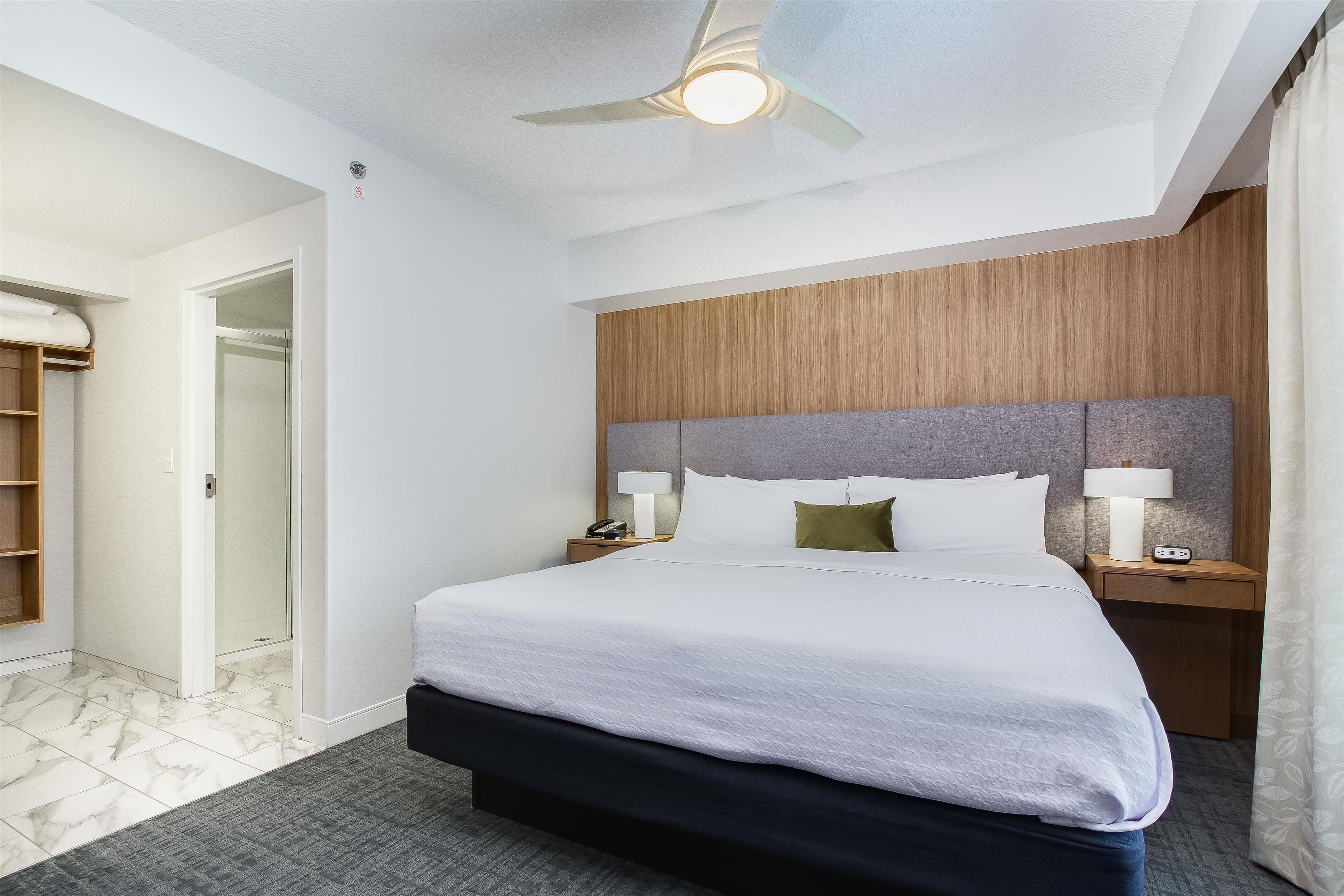 A hotel room with a large bed, wooden headboard, two bedside tables with lamps, a ceiling fan, and a bathroom with a glass shower door.