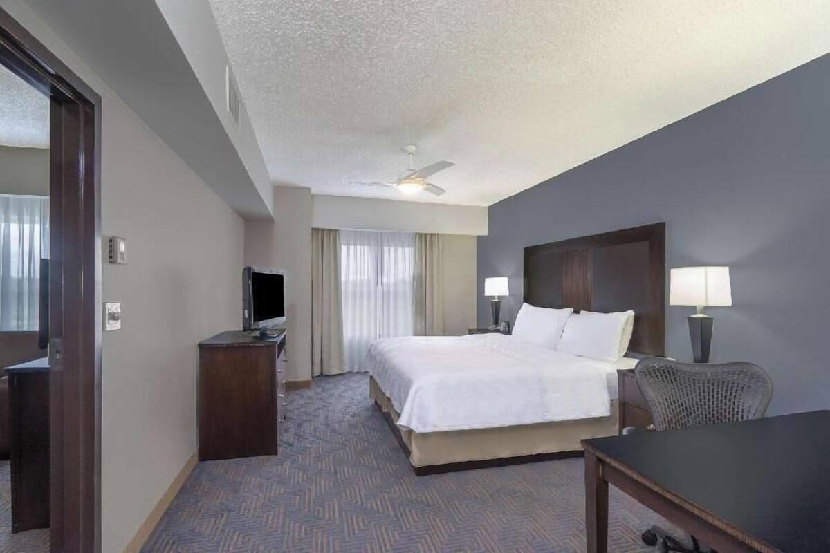 A hotel room with a bed, two bedside lamps, a desk, a chair, a television, and a ceiling fan.