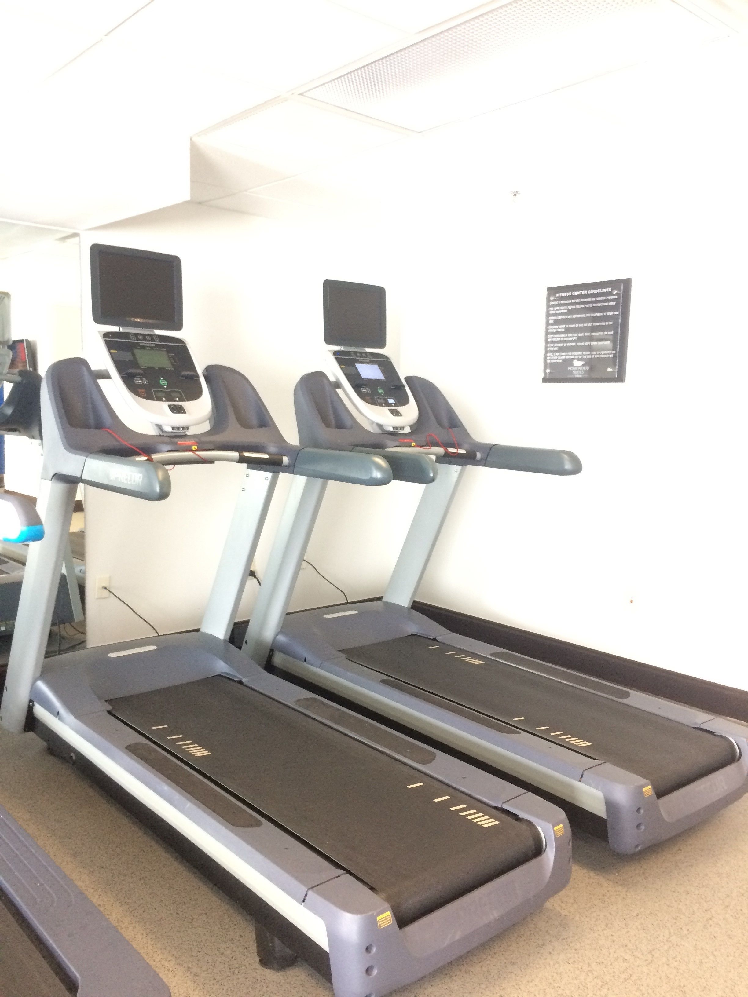 Three treadmills in a gym with a white wall and a sign about fitness center guidelines.